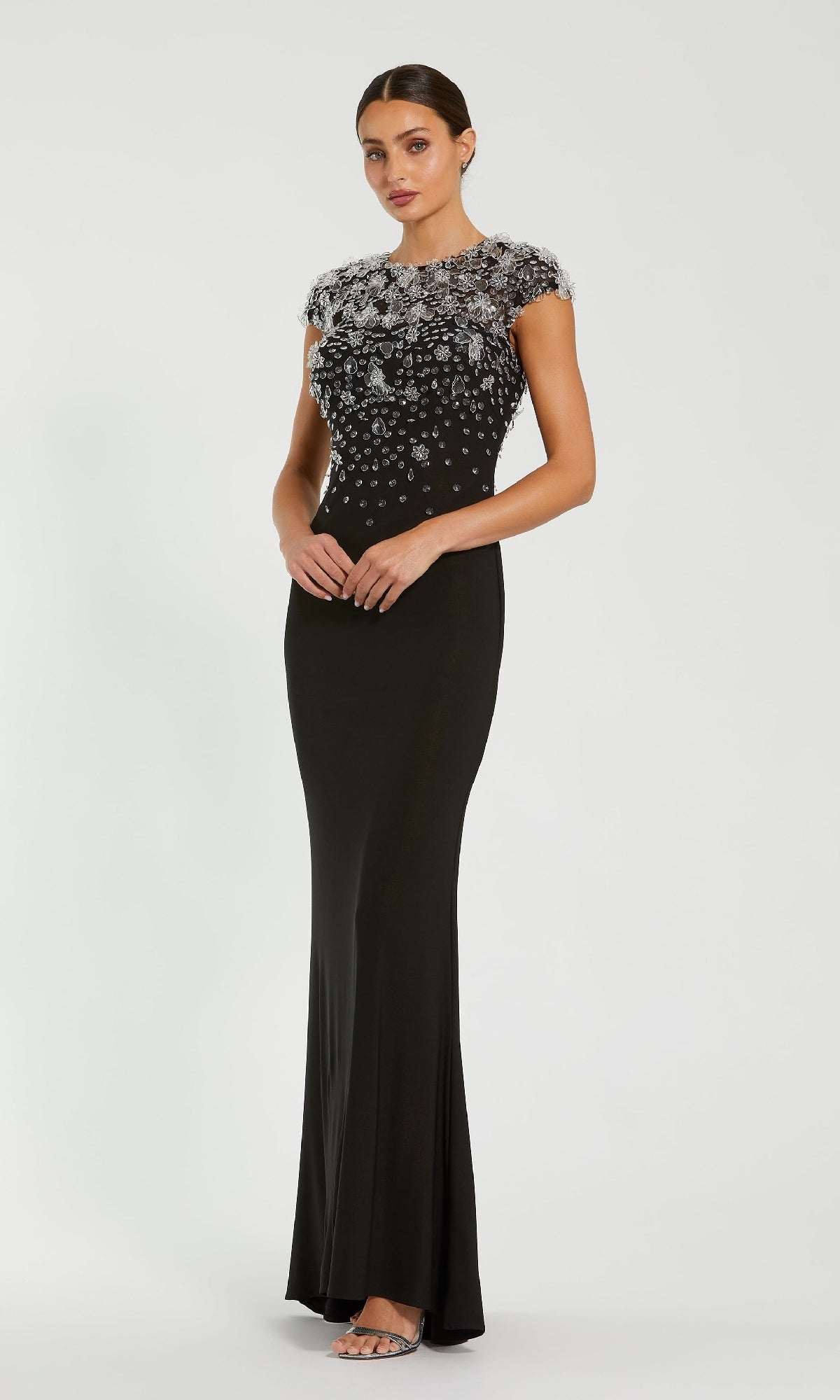 A woman models the Mac Duggal 44081 Cap Sleeve Black Formal Dress, featuring silver-embellished details on the bodice, standing against a plain white background.